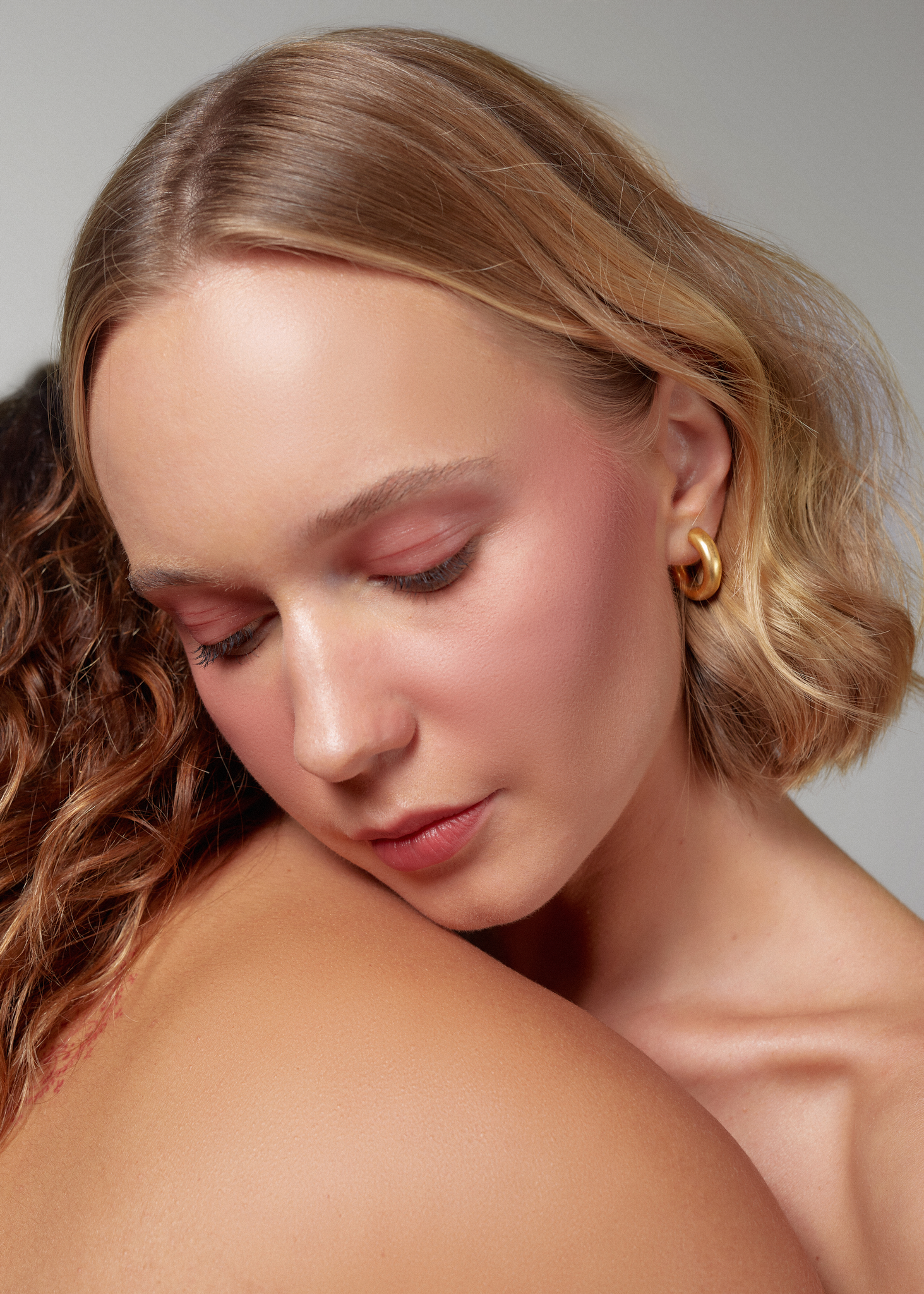 Portrait of a blonde woman with beautiful and glowy skin, leaning her head on the back of a brunette woman wearing boomii beauty's balms.